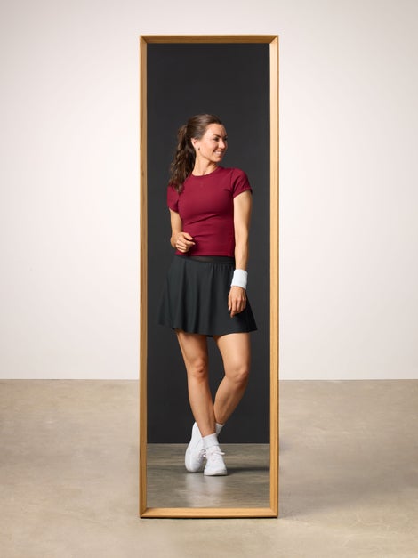 Woman reflected in mirror wearing a burgundy top and black tennis skirt. front view.