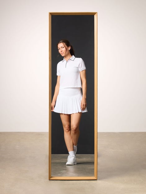 Woman reflected in mirror wearing white polo and white tennis skirt.