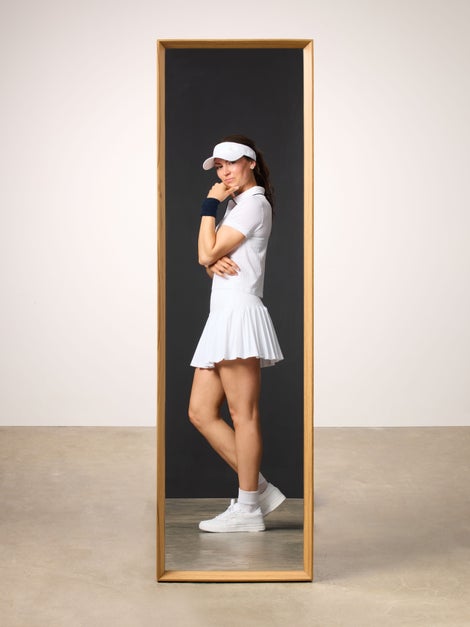 Woman reflected in mirror wearing white polo and white tennis skirt.