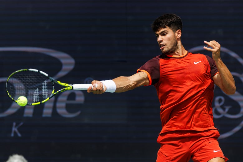 Carlos Alcaraz hits a forehand wearing a red and burgundy shirt and matching shorts.