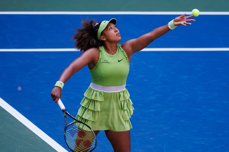 Naomi Oaska serves a tennis balll wearing a green ruffle tennis dress