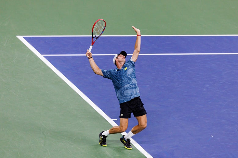Tommy Paul in mid-service motion wearing a New Balance polo in a blue print with navy shorts.