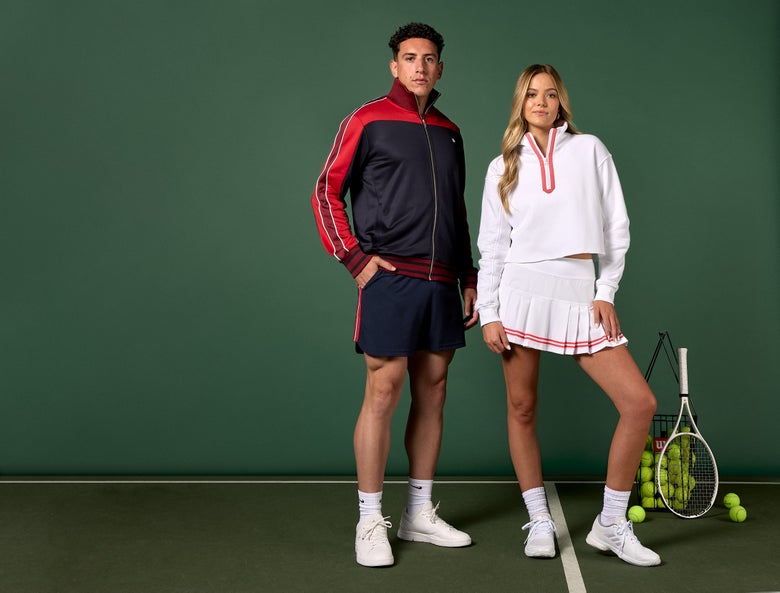 Man and woman wearing tennis jackets, shorts and skirt with basket of balls in background